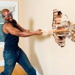 Un hombre con gafas de seguridad golpea con un mazo una pared de ladrillos para derribar la pared, causando daños visibles a la estructura.