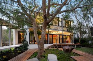 Casa moderna de dos pisos al atardecer con grandes ventanales, rodeada de árboles maduros y un jardín paisajístico con senderos de madera y asientos esculturales.