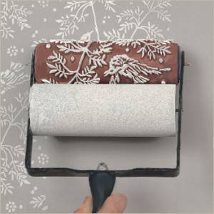 Un rodillo decorativo siendo utilizado para aplicar un diseño blanco floral y de pájaros en una superficie marrón.