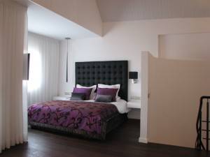 Dormitorio moderno con una cama grande con cabeceros para la cama únicos, ropa de cama morada y paredes en tonos neutros, situado junto a una escalera abierta sin barandilla visible.