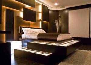 Un dormitorio moderno que cuenta con una cama elevada con iluminación tenue, tonos tierra y una pantalla de proyección en la pared iluminada por luz indirecta.