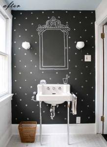La pared de un baño pintada de negro y decorada con un marco ornamentado dibujado con tiza, que incluye un lavabo con pedestal blanco, apliques de pared sencillos y una pequeña papelera de mimbre, se transforma en una pared