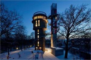 Una torre de agua iluminada de varios niveles y de diseño moderno destaca en medio del crepúsculo, rodeada de un paisaje nevado y árboles sin hojas.
