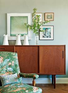 Un rincón decorado con buen gusto, que muestra un aparador de madera con un jarrón de flores blancas encima, acompañado de obras de arte enmarcadas en una pared de color verde pálido, reflejo del estilo nórdico.