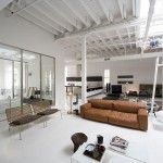 Un espacioso y moderno apartamento tipo loft convertido de un espacio comercial con paredes blancas, techos altos y una combinación de muebles clásicos y contemporáneos. Las características incluyen un plano de planta abierto, piso blanco pulido, un piso marrón