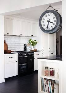 Una cocina moderna con gabinetes blancos, placa para salpicaduras de azulejos tipo metro y encimeras negras con un gran reloj de pared, un horno negro y una pequeña isla con espacio para guardar libros de cocina, diseñada en un estilo industrial.