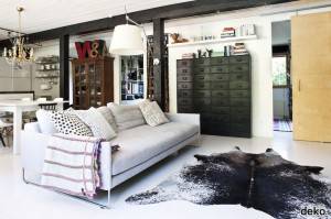 Una sala de estar moderna con un sofá gris con cojines estampados, una alfombra de piel de vaca negra sobre un piso blanco, un gran archivador de metal, una lámpara colgante blanca y estantes de madera con varios elementos decorativos.