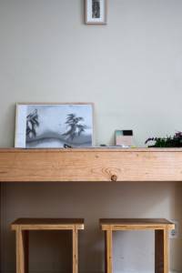 Una habitación minimalista con un escritorio de madera, dos taburetes a juego y una impresión artística sin marco apoyada contra la pared del escritorio muestra la estética japonesa.
