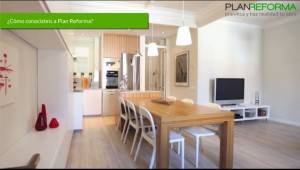 Un espacio de vida moderno y diáfano que cuenta con una zona de comedor con mesa y sillas de madera, una cocina con electrodomésticos al fondo y una zona de estar a la derecha con un televisor. Sobre el