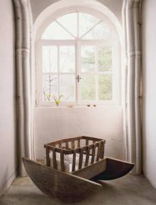 Una cuna de madera está volcada junto a una ventana arqueada con vista a los árboles, ubicada dentro de una casa sencilla con paredes y cortinas blancas.
