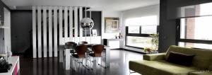 Una moderna sala de estar con paredes negras, que cuenta con un sofá verde, una zona de comedor con una mesa blanca y sillas marrones, y grandes ventilaciones.