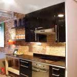 Una cocina moderna con gabinetes negros, electrodomésticos de acero inoxidable y una estantería de madera contra una pared de ladrillo visto muestra el diseño de Cuales.