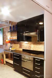 Una cocina moderna con gabinetes negros, electrodomésticos de acero inoxidable y una estantería de madera contra una pared de ladrillo visto muestra el diseño de Cuales.