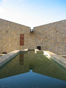 Edificio de piedra con una piscina reflectante bajo un cielo azul claro, diseñado por el arquitecto Toni Morlà para el concursoPR de arquitect