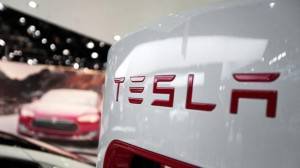 Primer plano de la parte trasera de un vehículo Tesla blanco con el logotipo de Tesla, con otro coche Tesla rojo con baterías de Tesla y un cartel promocional difuminado al fondo.