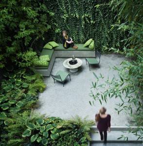 Una persona se relaja en un sofá verde al aire libre rodeada de exuberantes jardines verticales monocromáticos y plantas en macetas, mientras otra persona pasa.