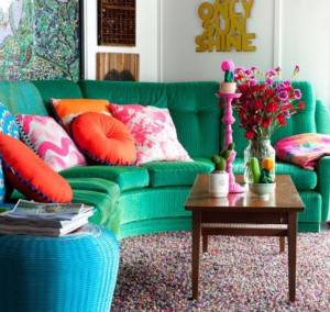 Una vibrante sala de estar con un sofá de textura verde adornado con coloridos cojines, una mesa de café de madera con un candelabro rosa y un jarrón de flores, frente a un piso cubierto de cuentas multicolores y una pared ecléctica.
