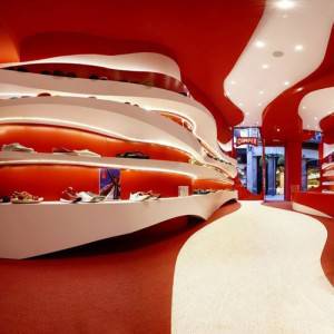 Interior moderno de una zapatería minorista con estantes ondulados en rojo y blanco con calzado en exhibición, acentuado por formas orgánicas y un camino curvo que atraviesa el espacio.