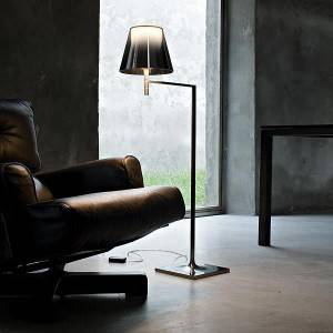 Una lámpara de pie moderna con un diseño de pantalla único se encuentra junto a un sillón de cuero oscuro en una habitación con paredes de concreto y una vista clara de la vegetación exterior, que encarna las cosas ideales para tener en