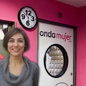 Una mujer sonríe en un espacio interior con paredes rosas, cerca de una puerta con el texto "ondamujer.com" y un reloj de pared que muestra la hora encima de ella. Al lado, hay