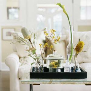 Una bandeja con una variedad de artículos decorativos para tener en casa, incluidos jarrones con flores secas, un plato de vidrio cubierto y botellas pequeñas, colocadas sobre una mesa en un espacio habitable en tonos claros.