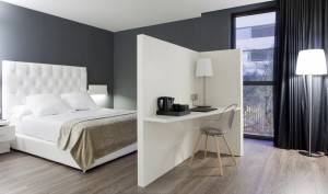 Una habitación moderna del Hotel SB Plaza Europa con una cama grande con cabecera blanca, paredes oscuras, un pequeño escritorio con una silla y una lámpara de pie cerca de la ventana.