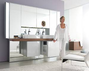Una persona con atuendo blanco caminando en un baño moderno diseñado para ganar espacio, con un lavabo doble y grandes espejos.
