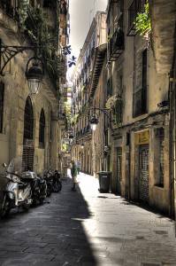 Una calle estrecha e iluminada por el sol en una ciudad histórica europea, flanqueada por edificios altos con balcones y adornada con plantas colgantes, mientras una persona solitaria camina por el camino adoquinado junto a motocicletas estacionadas.