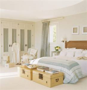 Un dormitorio cuidadosamente organizado, decorado piso por un interiorista, con una cama grande, adornada con ropa de cama blanca y azul claro, flanqueada por mesas auxiliares estilo equipaje y un cómodo sillón.