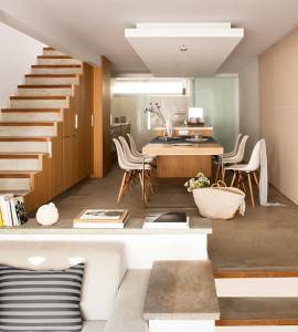 Una cocina moderna con elementos de madera y un comedor, diseñada por Cotacero Taller de Arquitectura, cuenta con una escalera flotante a la izquierda y un acogedor rincón para sentarse en el