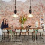 Una zona de comedor rústica que presenta una mesa y sillas de estilo industrial con un fondo de pared de ladrillos desgastados, complementada por lámparas col.
