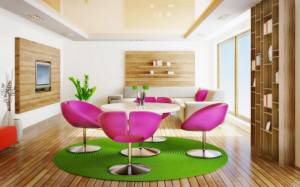 Comedor moderno decorado con sillas de color violeta brillante alrededor de una mesa blanca, colocada sobre una alfombra circular verde, con pisos y estanterías de madera, mucha luz natural y una decoración minimalista.