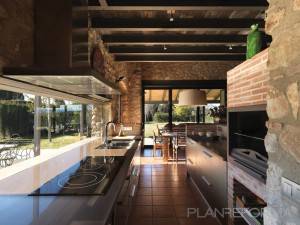 Moderna zona de cocina al aire libre con electrodomésticos integrados y elementos de diseño rústico, con paredes de piedra y ladrillo visto, debajo de un patio cubierto con vigas de madera con vistas al jardín. Ideal para quienes están considerando una "reforma cocina".