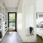 Un dormitorio estrecho y moderno con un armario empotrado blanco a la izquierda, una cama al final de la habitación y un espejo de cuerpo entero montado en la pared derecha, que refleja una vista de un