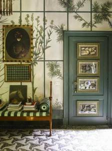 Una entrada de estilo vintage con papel tapiz botánico, una puerta verde oscuro adornada con arte enmarcado, un banco de madera con libros y un bastón decorativo.
