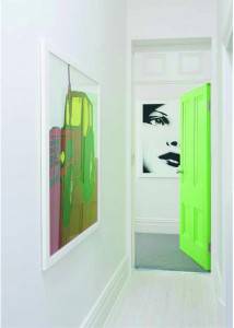 Una ideas casa: Un pasillo brillantemente iluminado con una puerta verde ligeramente entreabierta; en la pared izquierda cuelga una pintura abstracta,