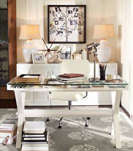 Una configuración de oficina en casa con un escritorio blanco con tapa de cristal, flanqueado por dos lámparas de mesa y una silla blanca tapizada. Sobre el escritorio hay material de oficina, libros,