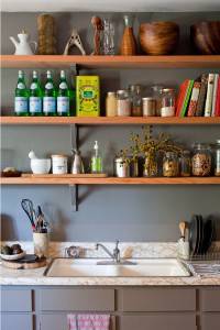 Una encimera de cocina ordenadamente organizada con un fregadero y repisas flotantes mostrando utensilios de cocina, botellas, frascos y libros de cocina.
