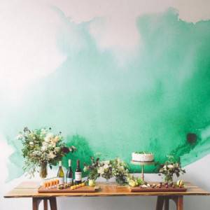Una mesa de madera con tendencia de decoración festiva, flores, un pastel y botellas en frente de una pared con una media pared de lavado acuarela verde.