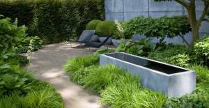 Un espacio de jardín moderno con un camino de grava, jardineras de concreto, exuberante vegetación ideal para dar sombra (jardines a la sombra) y un par de sillones al aire libre.