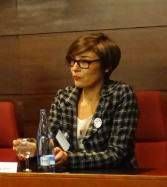 Una mujer con una chaqueta a cuadros y gafas sentada en una mesa con un micrófono y botellas de agua, que parece ser parte de un panel o discusión de Internet.