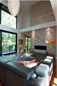 Salón moderno con techos altos, grandes ventanales, chimenea de piedra, muebles contemporáneos y abundante iluminación natural.