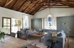 Una sala de estar decorada con gusto, con un techo de vigas de madera expuestas, armarios grises, asientos cómodos y una elegante decoración bajo la luz.