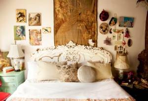 Un dormitorio de inspiración vintage con una cabecera blanca ornamentada, una variedad de almohadas decorativas y objetos y arte ecléctico en las paredes colocados sobre la cama.