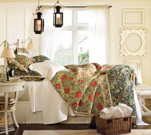 Un dormitorio amplio y luminoso con dos camas individuales con textiles florales verdes y rojos, complementadas con paredes blancas, linternas de estilo vintage y una canasta de toallas dobladas a los pies de una cama.