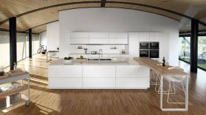 Una cocina moderna con gabinetes blancos y electrodomésticos, cuenta con una isla central, suelos de madera y una pared curva de vidrio que ofrece.