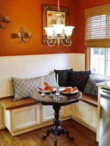 Un acogedor rincón comedor con una pared naranja, revestimiento de madera blanco y una mesa redonda de madera con una base de pedestal clásica. El área está adornada con candelabros blancos, apliques de pared.