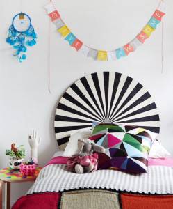 Un dormitorio colorido y ecléctico con una cabecera a rayas, una almohada con motivos geométricos, una colcha multicolor y un cartel decorativo que dice "Oliver es increíble" colgado en la pared. esta casa