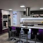 Una cocina moderna con gabinetes morados, electrodomésticos de acero inoxidable, encimera blanca, paneles de LED debajo de los gabinetes y barra desayunadora con taburetes.