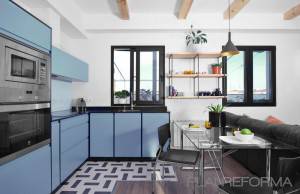 Cocina moderna con gabinetes azules, electrodomésticos de acero inoxidable y un pequeño comedor con iluminación colgante en un ático, que presenta una combinación de elementos de diseño industrial y contemporáneo.